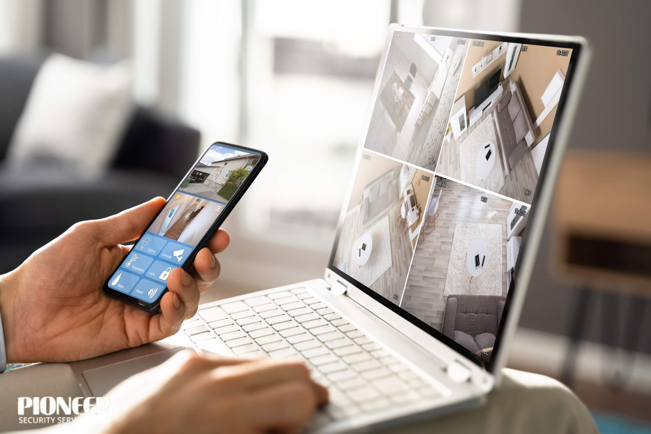 A person is monitoring home security on two devices in a bright living room. The laptop displays a four-panel grid of indoor surveillance feeds, while the person holds a smartphone showing a single outdoor camera feed along with smart home control buttons.