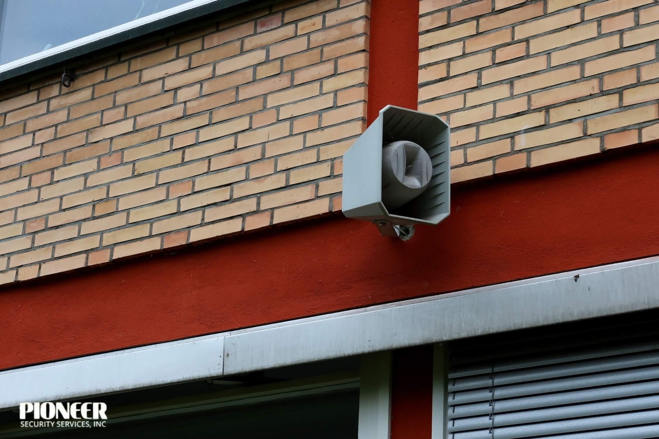Grey emergency or alarm siren speaker mounted on a light brown brick wall with a dark red painted concrete stripe below it, and a window awning visible at the bottom.