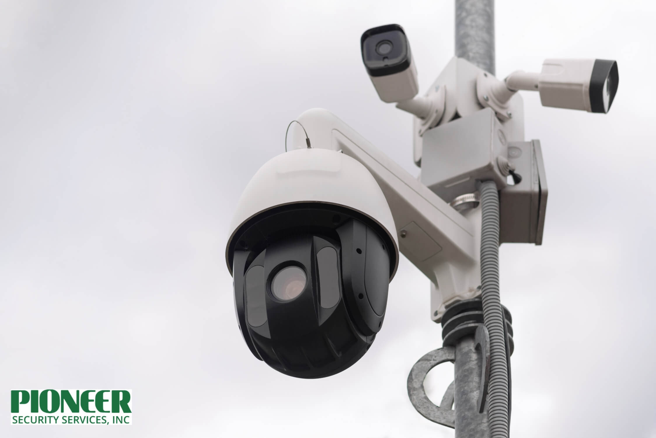 A close-up, low-angle shot of a multi-camera traffic monitoring system mounted on a pole against an overcast sky. It features a large, black, dome-shaped PTZ (Pan-Tilt-Zoom) camera and two smaller, white bullet-style cameras.