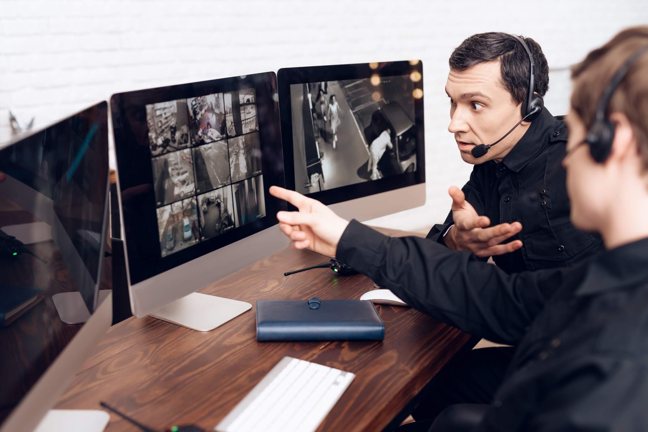 Two security guards in dark uniforms stand in a control room, looking at a large wall-mounted monitor that displays a six-panel grid of surveillance footage from interior locations. One guard is using a digital tablet.