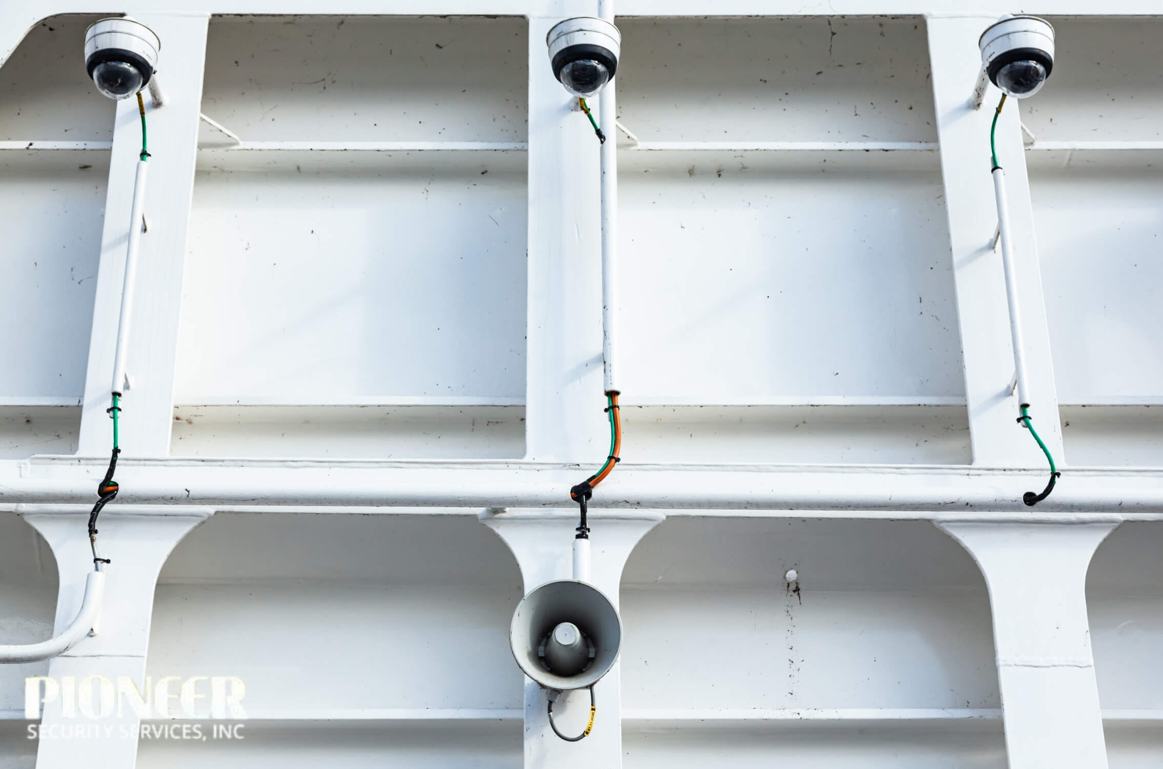 Symmetrical low-angle view of three dome-style security CCTV cameras and one horn loudspeaker mounted on the bright white, painted steel support beams of a ship or industrial structure.