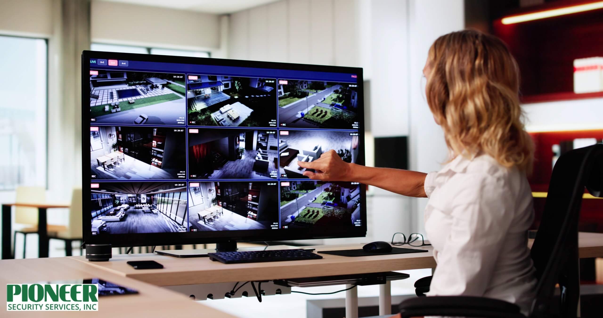 Woman sitting at an office desk, pointing to a large monitor displaying nine live security camera feeds showing interior and exterior views of a modern home or business property.