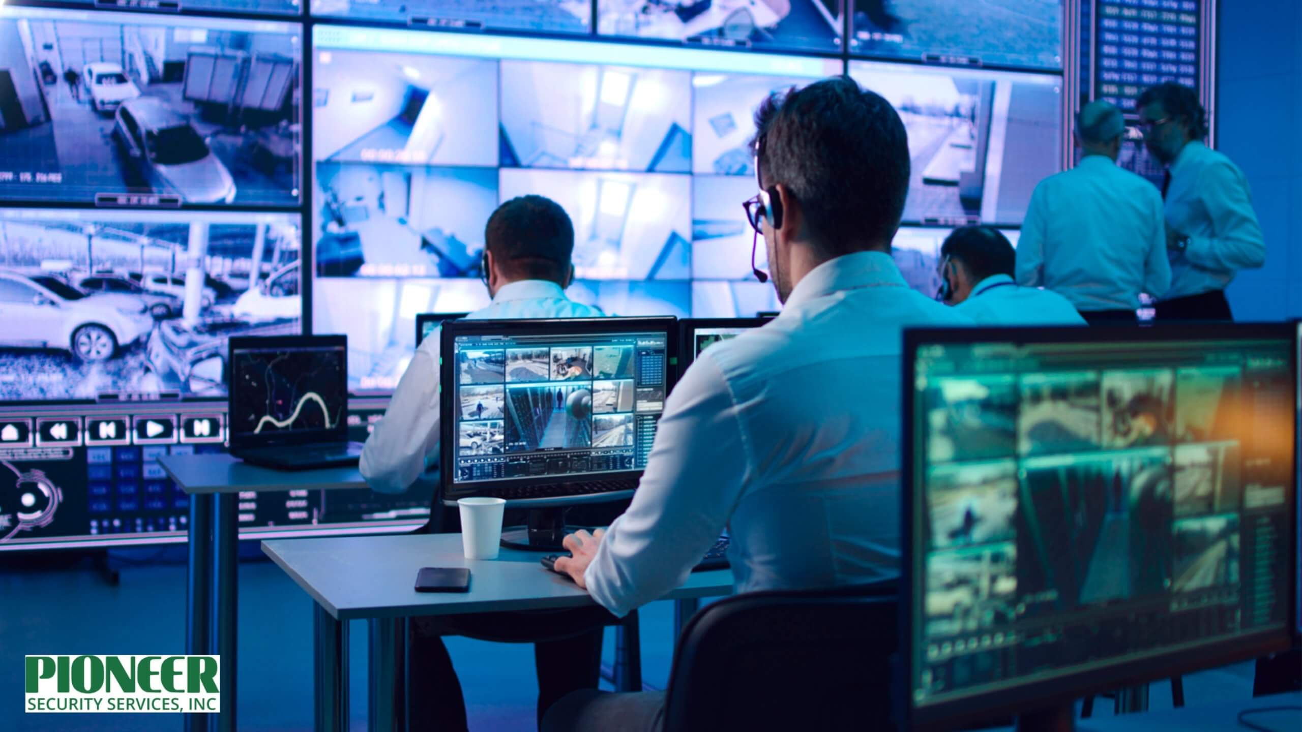 Security personnel working in a dimly lit surveillance control room. Several operators are seated at desks viewing multi-screen displays showing video feeds from various security cameras, with a large wall of screens displaying a grid of surveillance footage in the background.